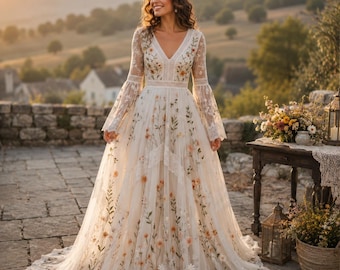 May include: A floor-length off-white dress featuring long bell sleeves, a V-neckline, and a fitted bodice. The fabric is adorned with embroidered colorful wildflowers and green stems throughout, with lace trim details at the waist and cuffs.