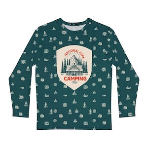 May include: A long-sleeved teal shirt featuring an all-over pattern of small white camping icons. Centered on the chest is a large cream-coloured shield emblem with a mountain and cabin graphic, reading "NATIONAL PARK CAMPING CLUB" in red text.