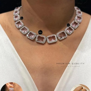 Puede incluir: Un collar con una serie de piedras rectangulares de color rosa claro, cada una rodeada por un borde de pequeñas piedras transparentes. El conjunto incluye pendientes rectangulares de color rosa claro y un anillo a juego, con monturas plateadas.