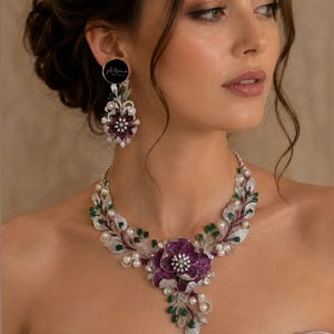 Könnte beinhalten: Ein Statement-Halsband und passende Ohrringe mit einem großen, zentralen, violetten Blumenmotiv, das mit Kristallen besetzt ist. Das Design umfasst weiße Perlen, kleine grüne tropfenförmige Steine und silberfarbene Metallblätter mit klaren Pavé-Kristallen.