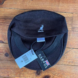 Pu&ograve; includere: Un berretto Kangol nero con finitura in maglia testurizzata e un logo a canguro azzurro ricamato sul davanti. Il berretto presenta una fascia interna, un'etichetta di manutenzione bianca e due cartellini di carta, uno dei quali indica la taglia M.