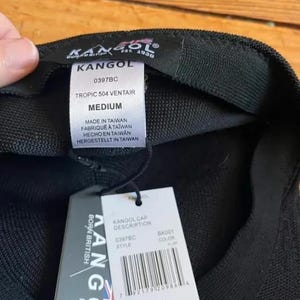 May include: A black Kangol Tropic 504 Ventair cap with a medium size label. The interior features a white tag with the text Kangol, 0397BC, Tropic 504 Ventair, Medium, and Made in Taiwan. A white retail hang tag with a barcode is attached by a black cord.