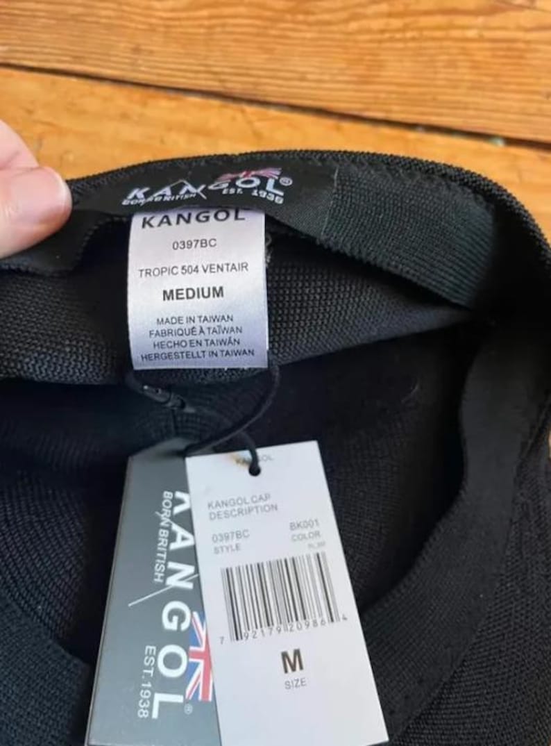 May include: A black Kangol Tropic 504 Ventair cap with a medium size label. The interior features a white tag with the text Kangol, 0397BC, Tropic 504 Ventair, Medium, and Made in Taiwan. A white retail hang tag with a barcode is attached by a black cord.