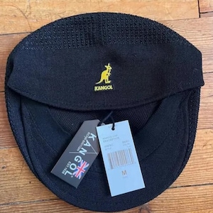 May include: A black Kangol cap with a textured knit finish and a yellow embroidered kangaroo logo. The cap features a front brim and two attached paper tags, one displaying the Kangol brand name, a Union Jack, and the text Born British Est. 1938.