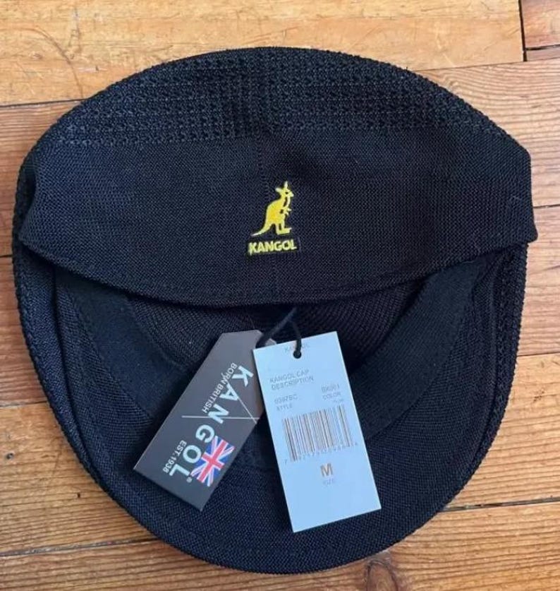 May include: A black Kangol cap with a textured knit finish and a yellow embroidered kangaroo logo. The cap features a front brim and two attached paper tags, one displaying the Kangol brand name, a Union Jack, and the text Born British Est. 1938.
