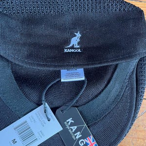 Pu&ograve; includere: Un berretto Kangol nero con una struttura in rete testurizzata. La parte anteriore presenta un logo a canguro bianco ricamato. Le etichette allegate mostrano il nome del marchio, il testo Born British, una bandiera britannica e la taglia M.