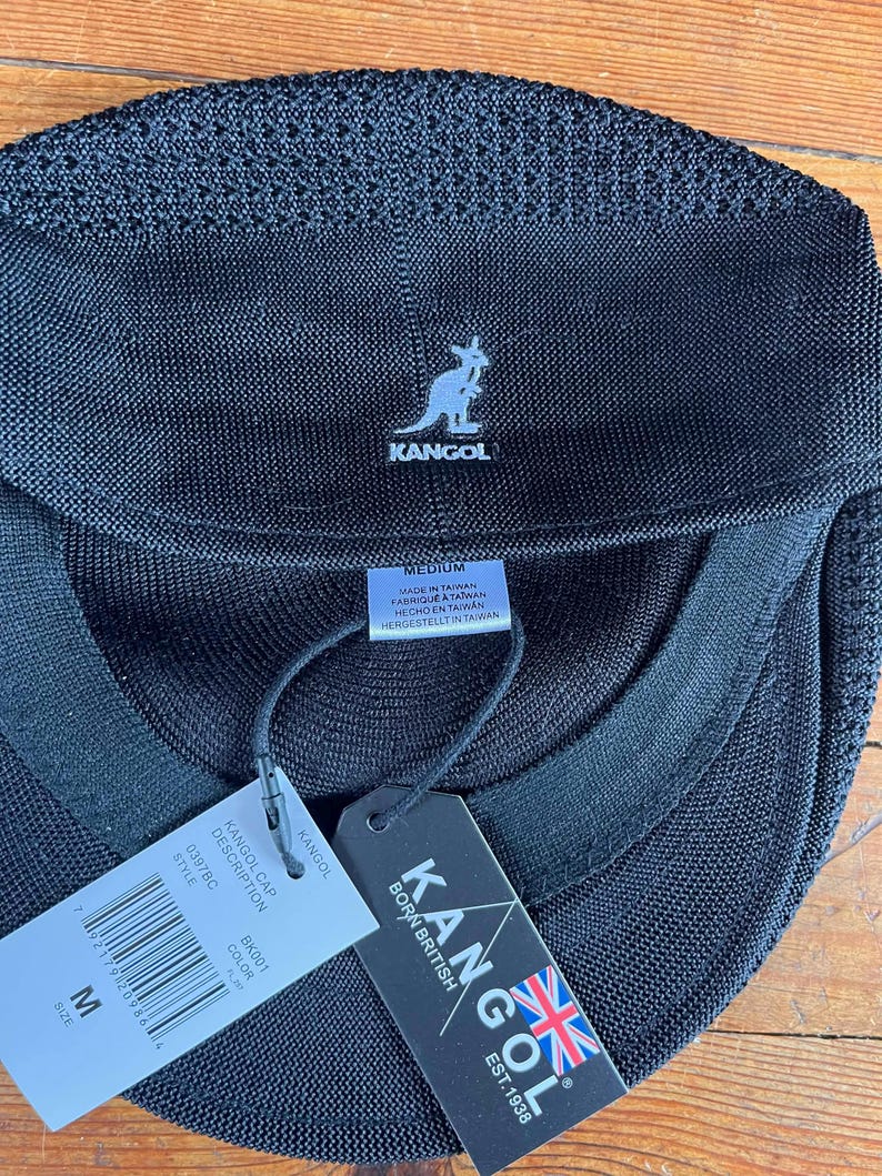 Pu&ograve; includere: Un berretto Kangol nero con una struttura in rete testurizzata. La parte anteriore presenta un logo a canguro bianco ricamato. Le etichette allegate mostrano il nome del marchio, il testo Born British, una bandiera britannica e la taglia M.