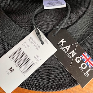 Pu&ograve; includere: Un berretto nero Kangol Tropic 504 Ventair con un'etichetta di taglia media. Le etichette allegate mostrano il logo Kangol, il testo Born British Est. 1938, un codice a barre, il numero di stile 0397BC, il codice colore BK001 e dettagli di produzione.