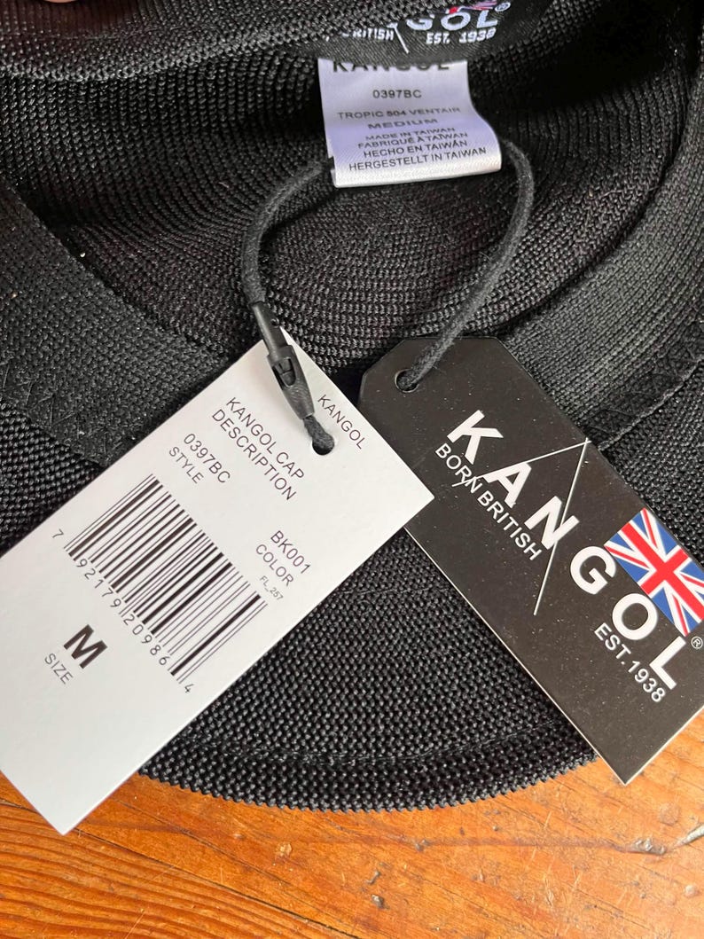 Pu&ograve; includere: Un berretto nero Kangol Tropic 504 Ventair con un'etichetta di taglia media. Le etichette allegate mostrano il logo Kangol, il testo Born British Est. 1938, un codice a barre, il numero di stile 0397BC, il codice colore BK001 e dettagli di produzione.