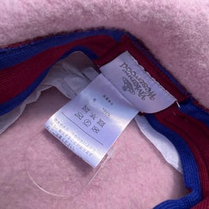 Puede incluir: Un sombrero de fieltro rosa claro con una etiqueta interior blanca que muestra el logotipo y el texto de Vivienne Westwood. La banda interior est&aacute; forrada con una tela a rayas azul brillante y rojo oscuro. Etiqueta de 100% lana.