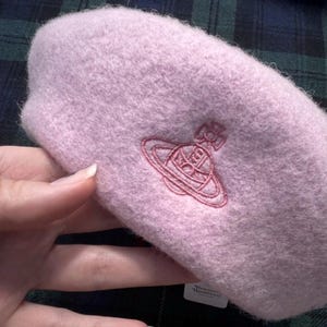 Puede incluir: Una boina suave y esponjosa de color rosa claro con un logotipo de orbe de Vivienne Westwood bordado en rosa m&aacute;s oscuro en la parte delantera. El gorro tiene una apariencia texturizada similar a la lana e incluye una etiqueta blanca.