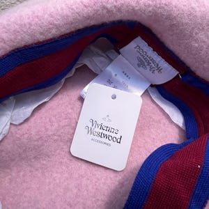 Puede incluir: Una boina de lana rosa claro con una banda interior a rayas de color rojo oscuro y azul. El interior incluye una etiqueta de marca blanca de Vivienne Westwood y una etiqueta de papel blanca con el texto Vivienne Westwood ACCESSORIES.