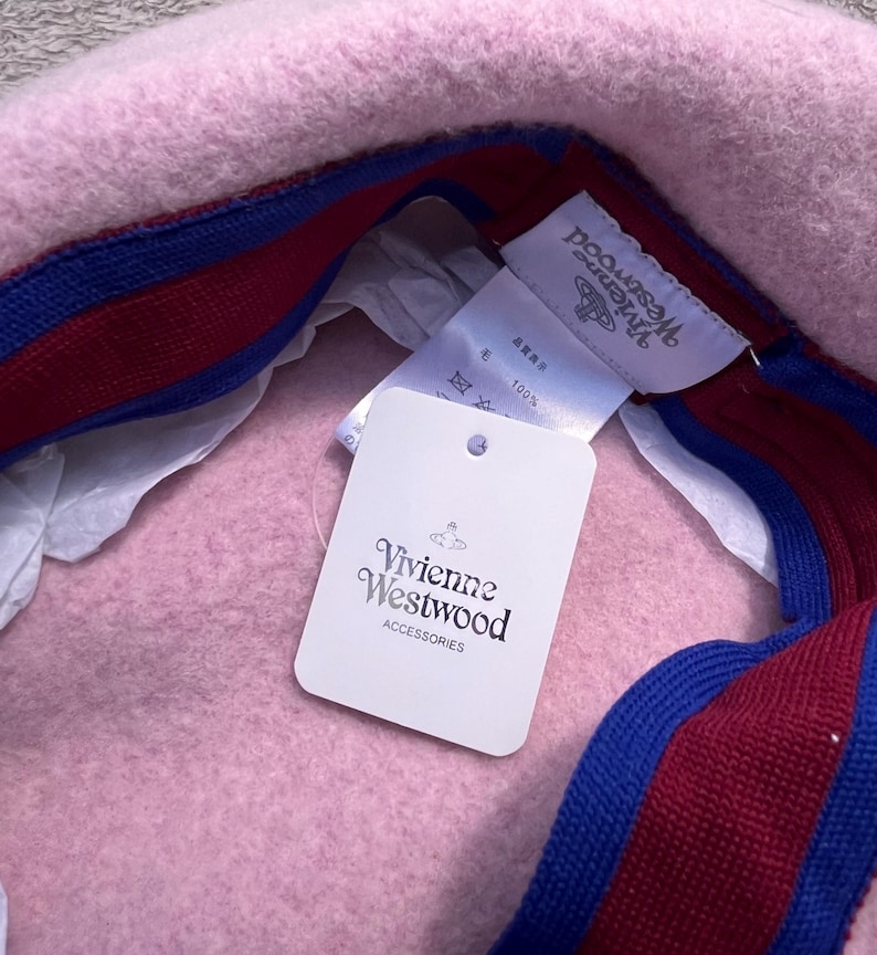Puede incluir: Una boina de lana rosa claro con una banda interior a rayas de color rojo oscuro y azul. El interior incluye una etiqueta de marca blanca de Vivienne Westwood y una etiqueta de papel blanca con el texto Vivienne Westwood ACCESSORIES.