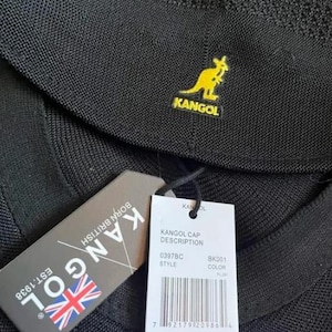 May include: A black textured cap featuring a yellow embroidered kangaroo logo and the text KANGOL. It includes a dark gray tag with the text KANGOL EST.1938 BORN BRITISH and a white tag with a barcode, size M, and the text KANGOL CAP DESCRIPTION.