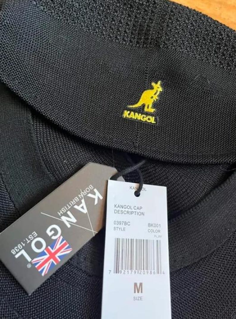 May include: A black textured cap featuring a yellow embroidered kangaroo logo and the text KANGOL. It includes a dark gray tag with the text KANGOL EST.1938 BORN BRITISH and a white tag with a barcode, size M, and the text KANGOL CAP DESCRIPTION.