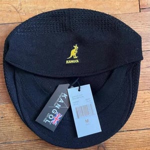 May include: A black Kangol cap with a textured knit finish and a yellow embroidered kangaroo logo. The cap features a front brim and includes two attached paper tags, one displaying the Kangol brand name, logo, and text, and the other showing size M.