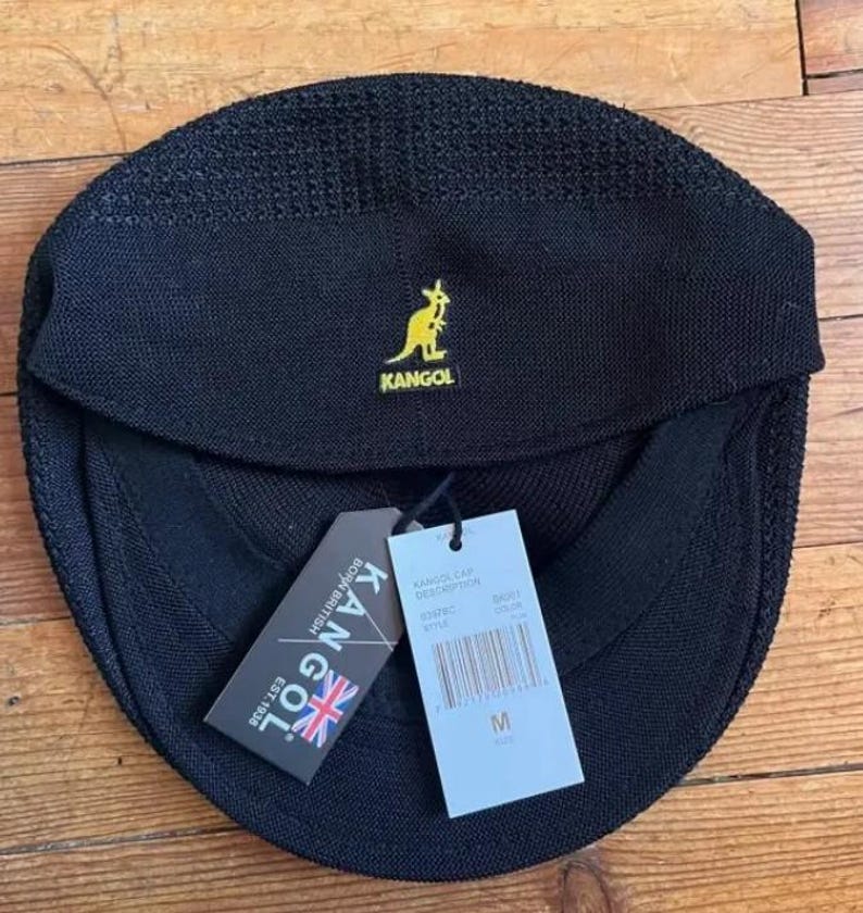May include: A black Kangol cap with a textured knit finish and a yellow embroidered kangaroo logo. The cap features a front brim and includes two attached paper tags, one displaying the Kangol brand name, logo, and text, and the other showing size M.