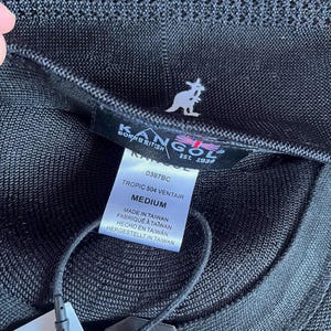 Pu&ograve; includere: Un berretto Kangol Tropic 504 Ventair nero in tessuto a rete testurizzato. L'etichetta interna mostra il logo Kangol, il testo Born British Est. 1938 e un'etichetta che indica la taglia Medium, modello 0397BC e Made in Taiwan con cartellino.