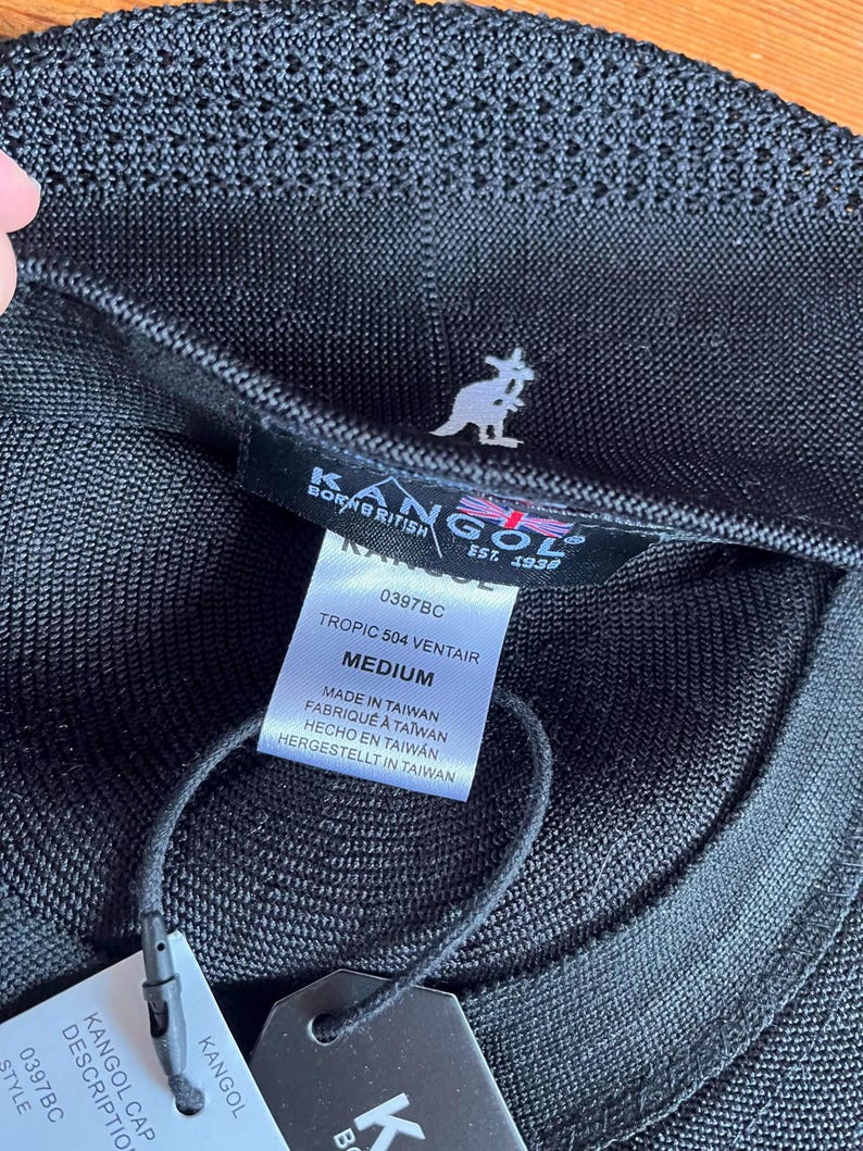 Pu&ograve; includere: Un berretto Kangol Tropic 504 Ventair nero in tessuto a rete testurizzato. L'etichetta interna mostra il logo Kangol, il testo Born British Est. 1938 e un'etichetta che indica la taglia Medium, modello 0397BC e Made in Taiwan con cartellino.