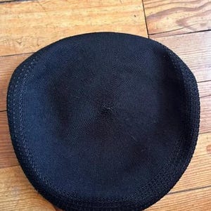 May include: A solid black beret featuring a circular knit construction with a subtle, textured pattern radiating from the center point to the outer edge. The hat has a defined, slightly thicker border along the circumference of the circular shape.
