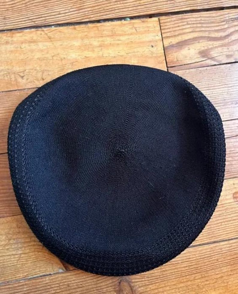 May include: A solid black beret featuring a circular knit construction with a subtle, textured pattern radiating from the center point to the outer edge. The hat has a defined, slightly thicker border along the circumference of the circular shape.
