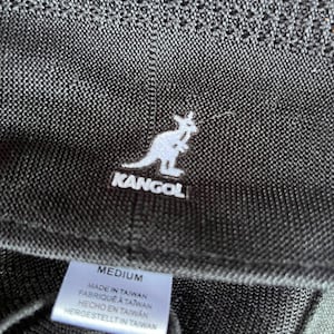Pu&ograve; includere: Un cappello a secchiello nero testurizzato con il logo del canguro Kangol ricamato in bianco. Un'etichetta interna bianca riporta la taglia MEDIUM e il testo MADE IN TAIWAN, FABRIQU&Eacute; &Agrave; TAIWAN, HECHO EN TAIWAN, HERGESTELLT IN TAIWAN in nero.