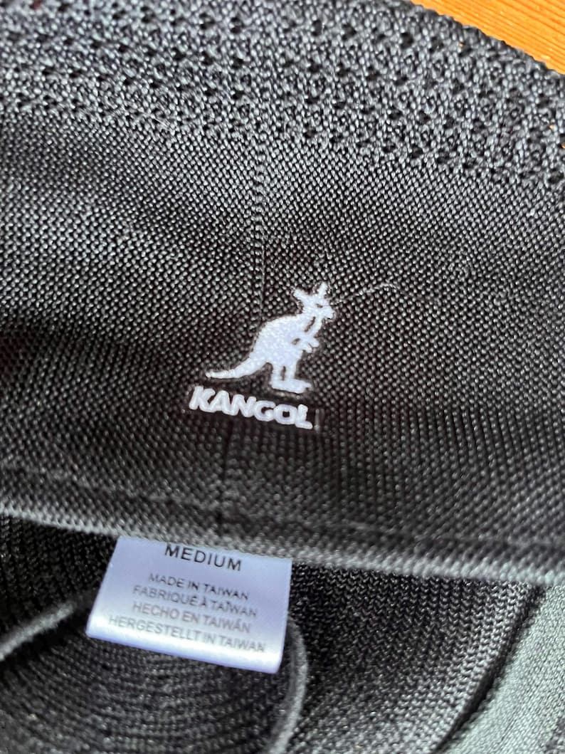 Pu&ograve; includere: Un cappello a secchiello nero testurizzato con il logo del canguro Kangol ricamato in bianco. Un'etichetta interna bianca riporta la taglia MEDIUM e il testo MADE IN TAIWAN, FABRIQU&Eacute; &Agrave; TAIWAN, HECHO EN TAIWAN, HERGESTELLT IN TAIWAN in nero.
