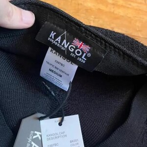 May include: A black Kangol Tropic 504 Ventair cap with a textured knit fabric. The interior features a black label with the Kangol logo and a British flag. A white tag displays the size Medium, style 0397BC, and manufacturing details in Taiwan.