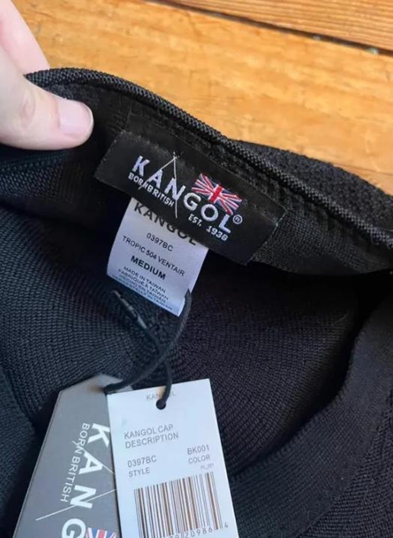 May include: A black Kangol Tropic 504 Ventair cap with a textured knit fabric. The interior features a black label with the Kangol logo and a British flag. A white tag displays the size Medium, style 0397BC, and manufacturing details in Taiwan.