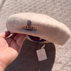May include: A beige wool beret featuring a dark brown embroidered orb logo on the front. The hat includes a blue and red interior lining and a white rectangular paper tag attached with a plastic fastener, displaying visible brand text.