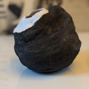 Może przedstawiać: Małe, zaokrąglone naczynie ceramiczne o wysoce teksturowanej, matowej czarnej powierzchni zewnętrznej. Górna część obiektu posiada gładki, kontrastujący biały szkliwiony otwór, tworząc wyraźne wizualne rozdzielenie pomiędzy dwoma kolorami.