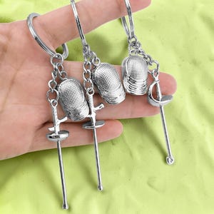 May include: Three silver-toned metal keychains, each featuring a miniature fencing mask charm and a small fencing foil sword charm attached to a circular split ring by a short silver chain link, showcasing detailed textures on the masks.