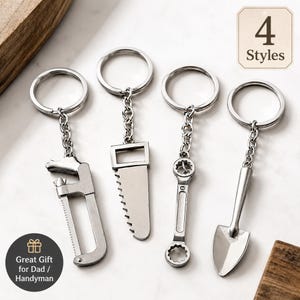 May include: Four silver-toned metal keychains featuring miniature tool charms: a hacksaw, a hand saw, a combination wrench, and a garden trowel. Each charm is attached to a circular split ring by a short metal chain. Text reads 4 Styles.