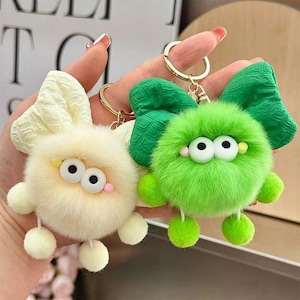 May include: Two fuzzy plush keychains, one cream and one bright green, each featuring a large textured fabric bow, two round white eyes with black pupils, small colored cheek accents, and two dangling plush feet attached to a gold metal ring.