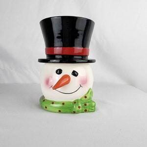 May include: A white ceramic snowman figurine with a black top hat, red trim, and a green and red polka dot scarf. The snowman has a carrot nose and black eyes.