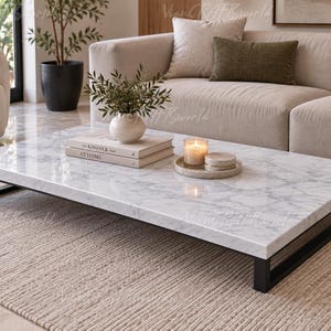 Pode incluir: Uma mesa de centro retangular com tampo de mármore branco com veios cinzentos e estrutura de metal preto. Sobre a mesa encontram-se um vaso branco com vegetação, dois livros empilhados intitulados Kinfolk e At Home e uma vela num tabuleiro.