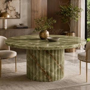 May include: A round dining table featuring a polished green onyx stone top with natural veining and a cylindrical, fluted base made of the same green stone. The table is surrounded by two beige upholstered chairs with thin gold metal legs.