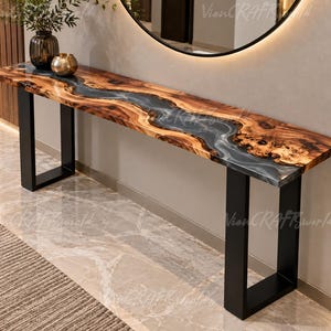 May include: A long console table featuring a natural live-edge wood slab with a central river-like inlay of translucent grey resin. The table is supported by two sturdy, rectangular black metal legs and displays a polished, smooth surface finish.