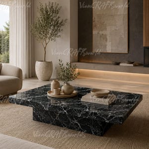 May include: A low-profile, rectangular coffee table featuring a polished black marble surface with prominent white veining. The table has a thick, block-style base and sits on a textured beige rug, topped with a wooden tray, ceramic vases, and books.
