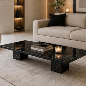 May include: A low-profile rectangular coffee table made of black marble with white veining. The table features a flat top surface supported by two thick, block-style legs. A stack of two books and a lit candle in a glass holder sit on the table.