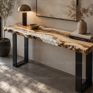 Puede incluir: Una mesa consola rectangular con una superficie de madera natural de borde vivo y una incrustación central de resina blanca. La mesa está sostenida por dos patas de metal negro en forma de U. La veta de la madera es visible y los bordes son rústicos.
