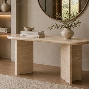 May include: A beige travertine console table with a smooth, oval-shaped top and two thick, rectangular slab legs. The natural stone surface features a porous, textured pattern in light tan and cream tones, providing a sturdy base for decor.