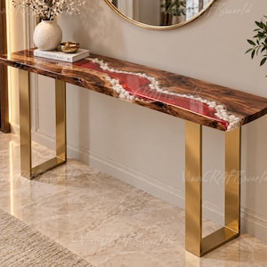 Puede incluir: Una mesa consola rectangular con un tablero de madera de borde natural que presenta una incrustación central de resina roja translúcida y piedras de río blancas. La mesa se apoya sobre dos patas de metal dorado cepillado en forma de U.