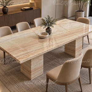 Puede incluir: Una mesa de comedor rectangular con una superficie de piedra pulida en tonos beige y crema con vetas lineales naturales. La mesa se apoya sobre dos bases de pedestal gruesas de piedra a juego y está rodeada por seis sillas tapizadas.