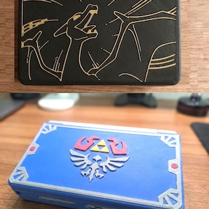 Può includere: Due custodie per console di gioco portatili. La custodia superiore è nera con un disegno a linee dorate di un drago. La custodia inferiore è blu con un emblema argento e rosso, un triangolo giallo centrale, dettagli angolari argento e punti rossi.