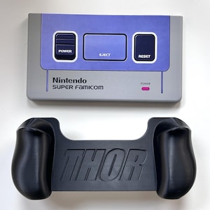 Könnte beinhalten: Eine graue und violette Nintendo Super Famicom Konsolen-Oberseite mit Power-, Eject- und Reset-Tasten sowie ein schwarzer Controller-Griff mit dem eingeprägten Wort THOR in der Mitte, beides auf einer schlichten weißen Oberfläche.