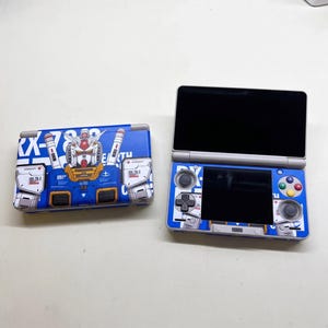 Op de afbeelding: Een draagbare spelcomputer met een blauw-witte afbeelding van de RX-78-2 Gundam-robot. Het apparaat wordt zowel gesloten als geopend getoond, met een dubbel scherm, een richtingspad, analoge sticks en vier gekleurde actieknoppen.