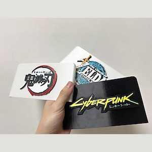 Könnte beinhalten: Drei rechteckige Kunststoffabdeckungen mit erhabenen Logos. Eine weiße Abdeckung zeigt das Demon Slayer-Emblem, eine weitere weiße Abdeckung das The Legend of Zelda-Logo und eine schwarze Abdeckung das gelbe Cyberpunk 2077-Grafiklogo.