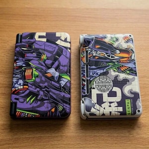 May include: Two rectangular handheld gaming consoles featuring custom skins. The left console displays a purple and green mechanical robot graphic. The right console features a white, purple, and black design with a circular speaker grille and the text SENPAI.