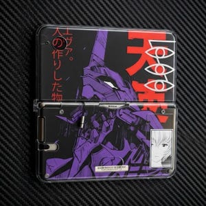 May include: A handheld gaming console featuring a purple mechanical robot illustration on a black background with red Japanese text, a white eye symbol, and a small rectangular portrait of a character with short hair on the lower right corner.